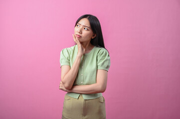 Asian woman thinking with hand on chin, looking up thoughtfully while standing against a plain pink...