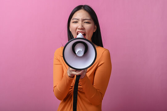Asian woman with long black hair wearing an orange dress holding a megaphone directly facing the camera, standing against pink background, symbolizing voice, announcement, or call to action