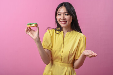 Asian woman in a yellow dress smiling while looking at a green-sprinkled donut in her hand, posing...