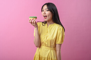 Asian woman in a yellow dress about to bite a green-sprinkled donut while looking at the camera,...