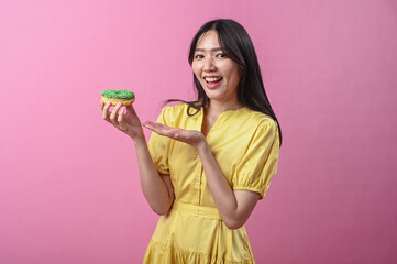 Asian woman in yellow dress smiling while holding a green-sprinkled donut and presenting it with...