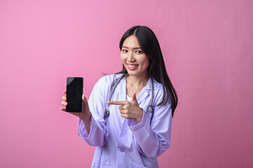 Asian female doctor smiling while holding and pointing at a smartphone with a blank screen,...