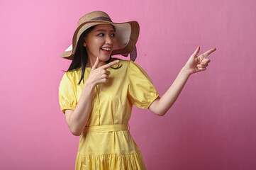 Asian woman in a yellow dress and sun hat smiling while pointing to the side with both hands,...