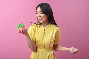 Asian woman in a yellow dress smiling while looking at a green-sprinkled donut in her hand, posing...