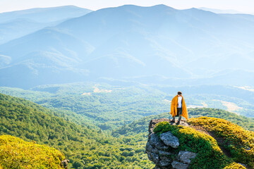 Man wrapped in a blanket standing on mountain cliff, admiring panoramic view of forest valley and...