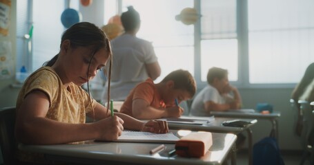 Elementary School Students Writing Exam or Doing Tasks in Notebooks. Female Teacher Walking Between...