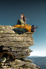 Young woman camping in mountains, sitting in sleeping bag on cliff at sunrise, enjoying peaceful...
