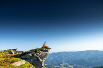 Man in yellow sleeping bag sitting on cliff edge, looking out over mountain landscape under clear sky