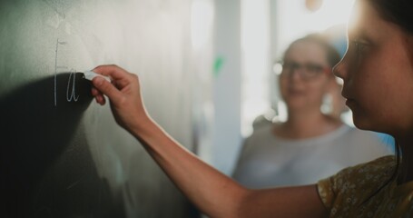 Close Up Shot of Primary School Girl Writing Word on the Board with Chalk. Female English Teacher Standing Nearby, Checking Correct Answer and Homework. English Grammar Lesson in Modern Classroom.