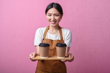 Asian woman in a brown apron smiling while holding a tray with two takeaway coffee cups, standing...