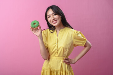 Asian woman in a yellow dress smiling while holding a green-sprinkled donut in front of a pink...
