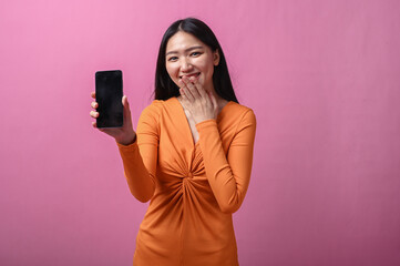 Asian woman with long black hair in orange dress standing indoors against pink background, holding...