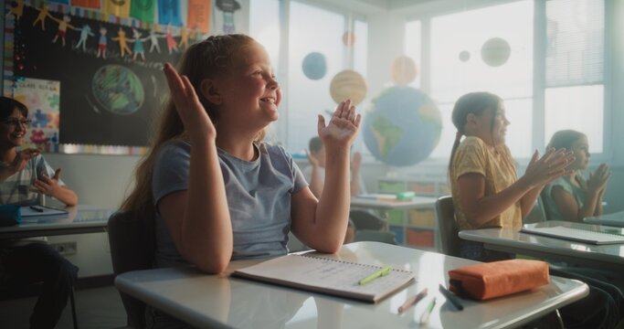 Female Teacher Conducting Funny Interactive Lesson to Primary School Children. Happy Young Boys and Girls Sitting at Desks, Raising Hands, Interacting with Teacher During Dynamic Classroom Activity.