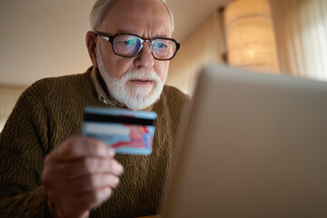 Elderly man with glasses looking at laptop holding a credit card with a concerned expression on his face