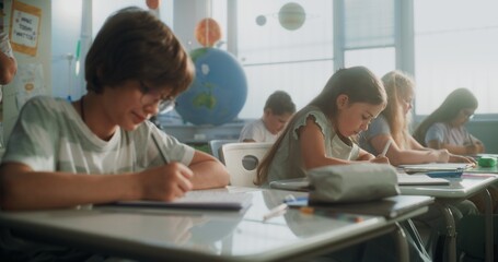 Elementary School Students Writing Exam or Doing Tasks in Notebooks. Female Teacher Walking Between...