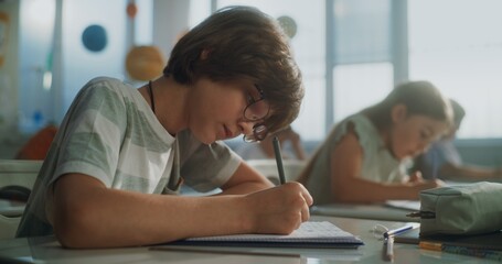 Primary School Boy Writing School Test, Doing Task in Notebook. Female Teacher Walking Between Desks, Controlling Group of Diverse Kids During the Exam Process in Modern Classroom. Close Up Shot.