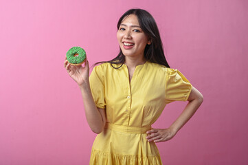 Asian woman in a yellow dress smiling while holding a green-sprinkled donut in front of a pink...