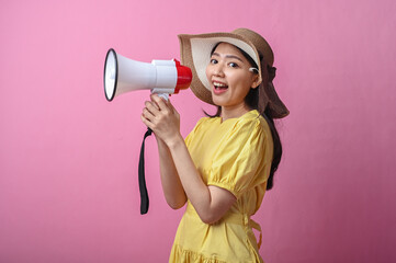 Asian woman in yellow dress and sun hat cheerfully shouting into a megaphone, standing against a...
