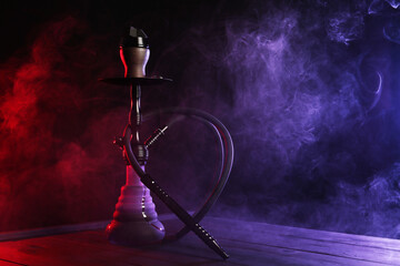 Hookah on wooden table with smoke in color lights against black background