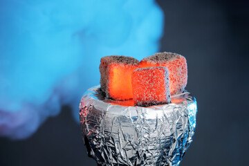 Smoldering charcoal cubes on hookah holder covered with foil and smoke in color light against dark...