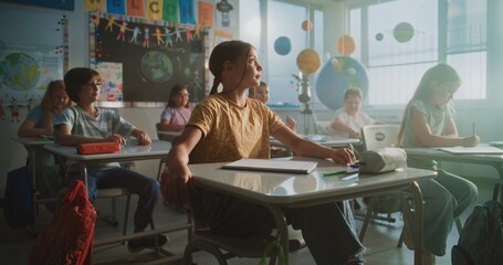 Primary School Children Sitting at the Desks, Listening to Lecture from Teacher, Raising Hands to...