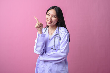 Smiling Asian female doctor in a white coat pointing upward with index finger, holding stethoscope,...