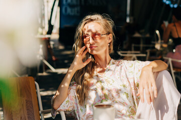 A beautiful blonde woman with long wavy hair sits on the terrace of a cafe on a summer day.