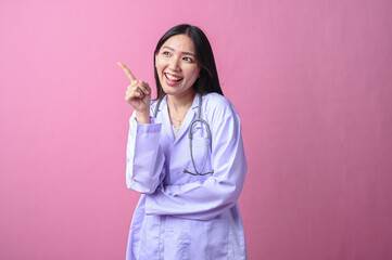 Smiling Asian female doctor in a white coat pointing upward with index finger, holding stethoscope,...