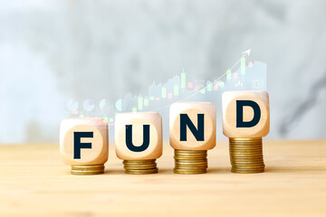 Wooden blocks spelling "FUND" on stacks of coins, with an ascending stock market chart in the background, symbolizing financial growth and successful investment.