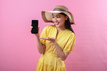 Asian woman in yellow dress and summer hat smiling while showing smartphone with one hand and...