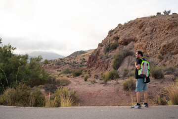 Latin man stands on a trail, pausing to enjoy the view of rocky hills covered with dry shrubs. The...