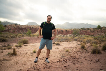 A man stands confidently with hands on hips amidst a vast, rugged desert terrain. Overcast skies...