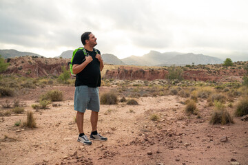Hispanic man stands in a dry, rocky landscape, gazing upwards. He wears a backpack and enjoys the...