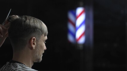 Professional barber grooming male client's hair with scissors, vintage barber pole framing...