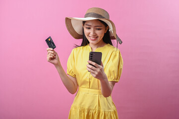 Asian woman wearing a summer hat and yellow dress smiling while holding a credit card and...