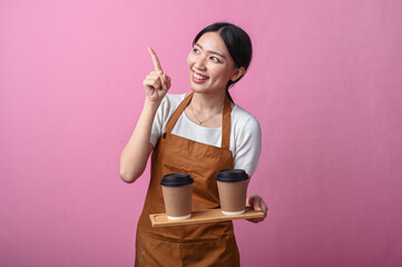 Asian woman in brown apron smiling and pointing up while holding a tray with two takeaway coffee...
