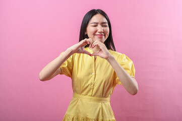 Asian woman in a yellow dress forms a heart shape with her hands while smiling warmly. She stands...