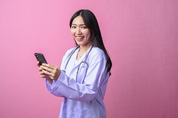Asian female doctor using a smartphone with both hands while smiling, dressed in a lab coat and...