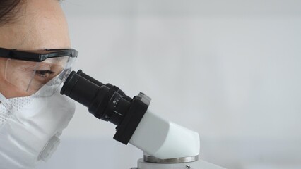 Close up of woman scientist wearing protective mask and goggles carefully examining sample through...