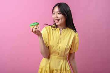 Asian woman in a yellow dress smiling while looking at a green-sprinkled donut in her hand, posing...