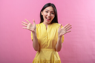 Asian woman in a yellow dress smiling brightly with both hands raised in an open wave gesture,...