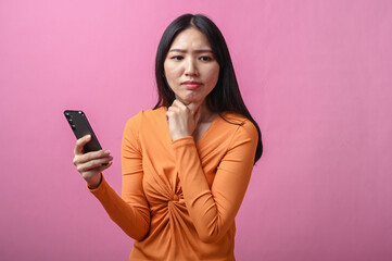 Asian woman with long black hair wearing orange dress holding smartphone and looking confused with...