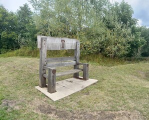 Rustic wooden bench with heart-shaped details and a metal letter &ldquo;T&rdquo; on the backrest. Set in a grassy clearing with trees in the background. A charming handcrafted seat in a natural setting.