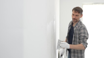 Construction worker wearing gloves and using a putty knife to plaster a white wall, performing home...