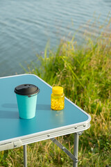 Blue portable camping table with a reusable cup and a pill bottle by the lake.