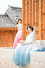 A Korean woman in her twenties wearing a sky-blue hanbok sits in front of the traditional facade of...