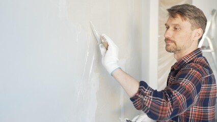 Male construction worker spreading plaster on white wall, wearing protective gloves during home...