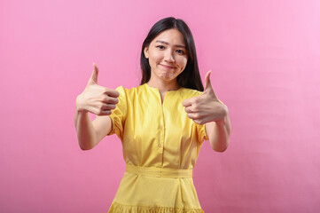 Asian woman in a yellow dress smiling with both thumbs up against a bright pink background....