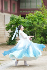 A Korean woman in her twenties wearing a sky-blue hanbok twirls gracefully in front of the...