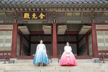Two Korean women in their twenties wearing pink and sky-blue hanbok stand in front of the facade of...